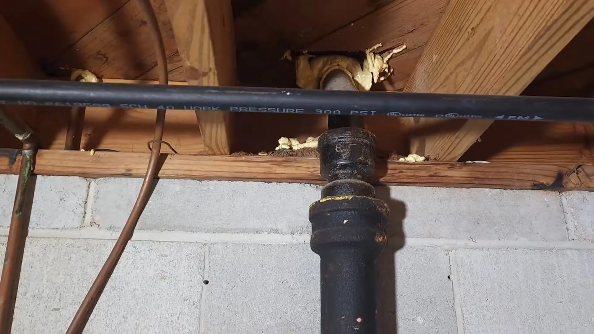 A black cast iron drain pipe, a copper line and a black supply pipe running through the wood joists of a basement ceiling
