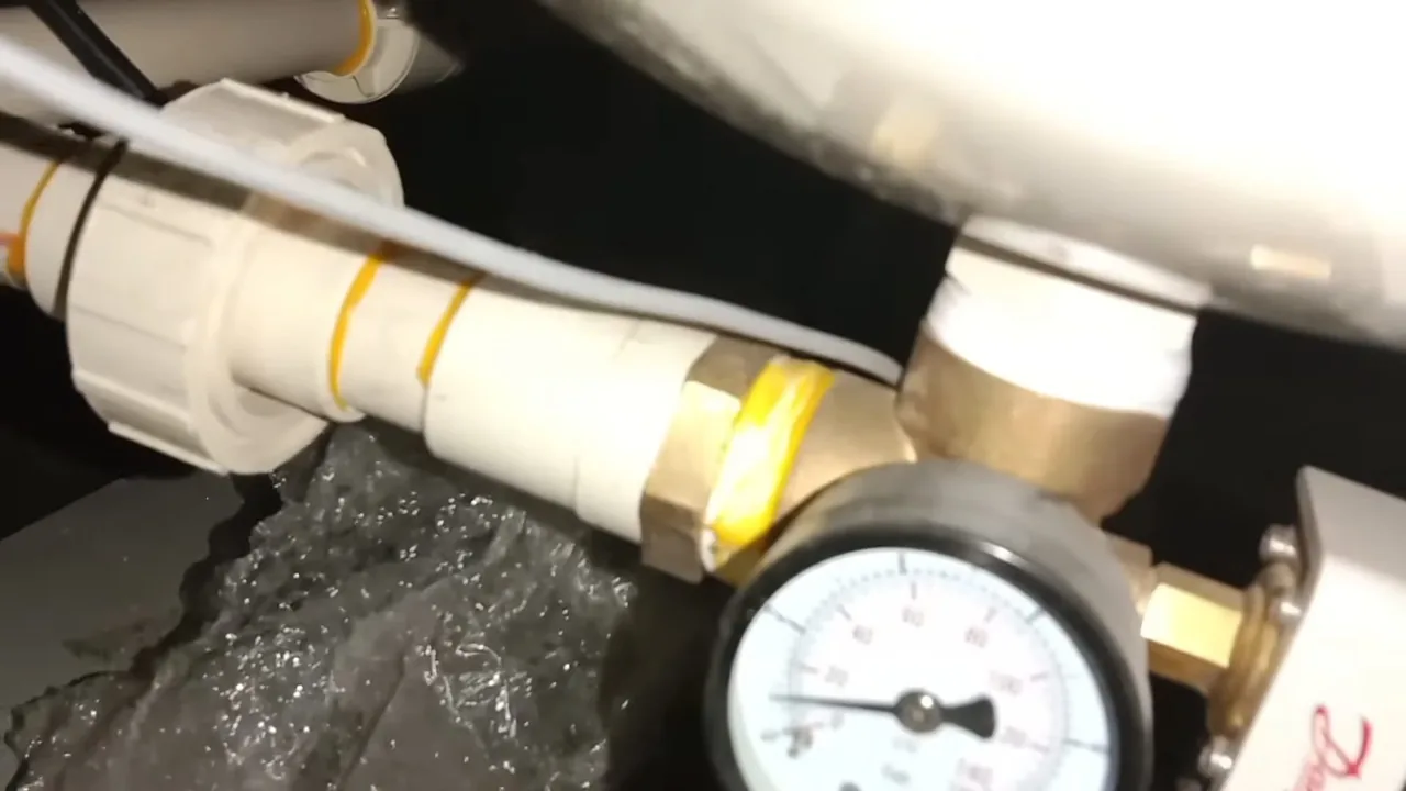 A brass pressure regulator with a round gauge attached to white pipe fittings with water spray below