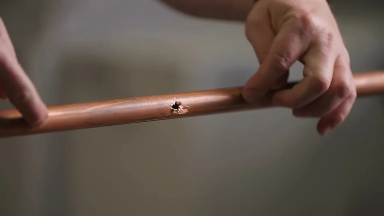 Two hands holding a length of copper pipe with a small pinhole puncture in its side