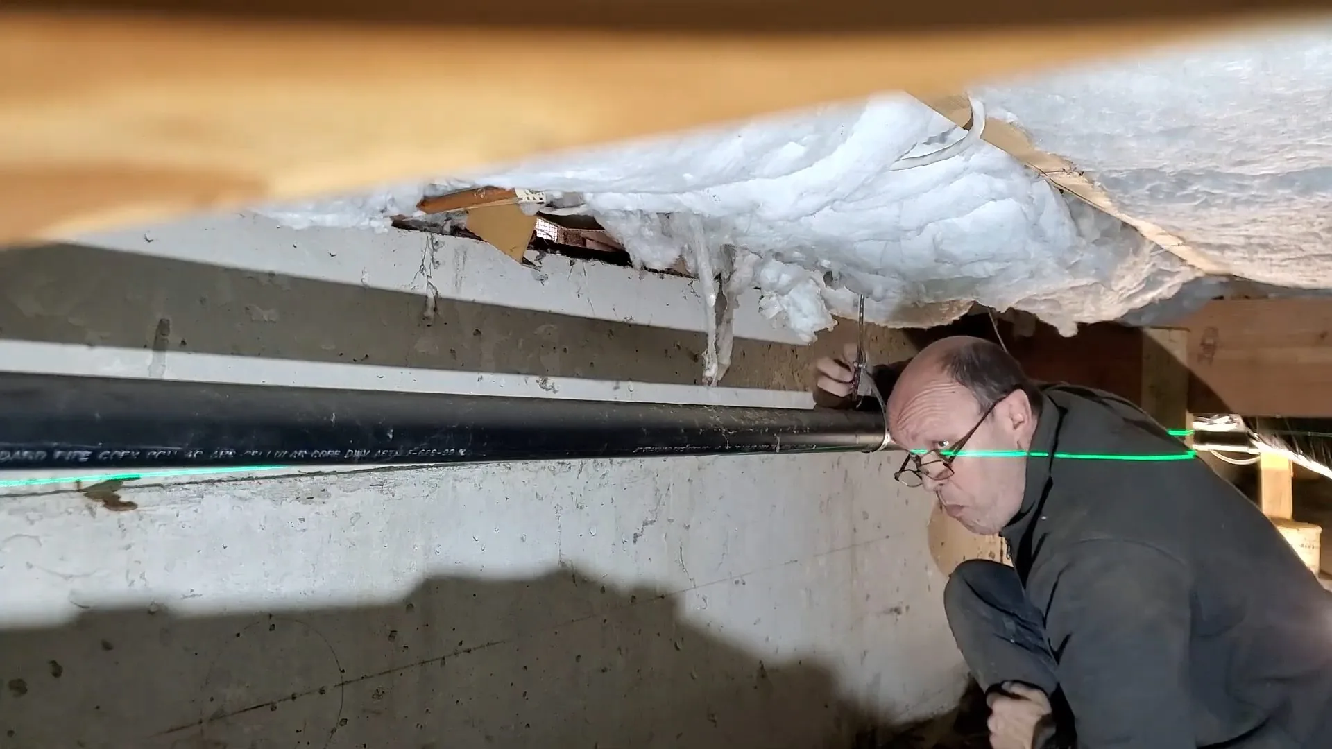 Man crouched in a crawlspace beside a long black pipe, with a green laser line across the frame