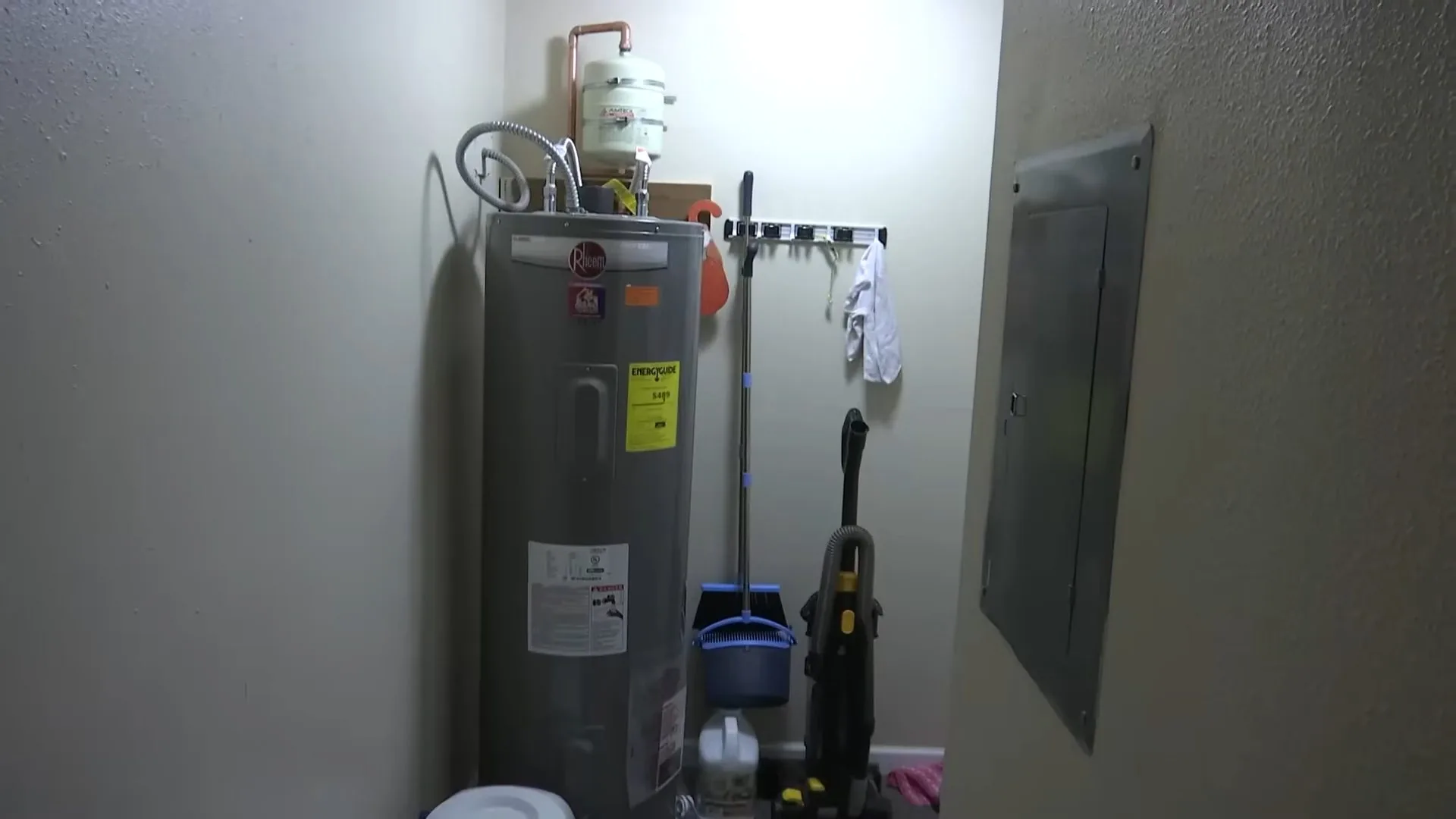 A tall gray tank water heater standing in a utility room beside an electrical panel cover, a broom and a dustpan