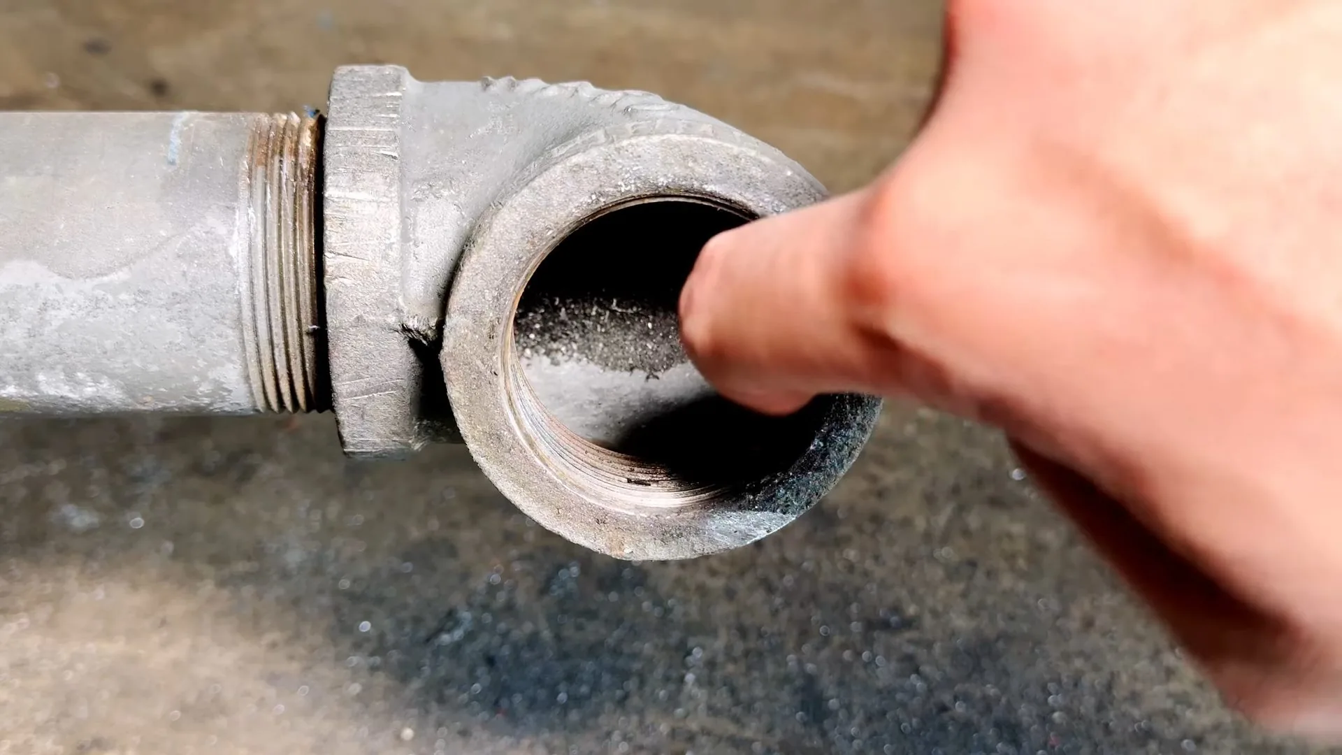 A finger touching the inside of a threaded cast iron cleanout opening on a gray pipe elbow