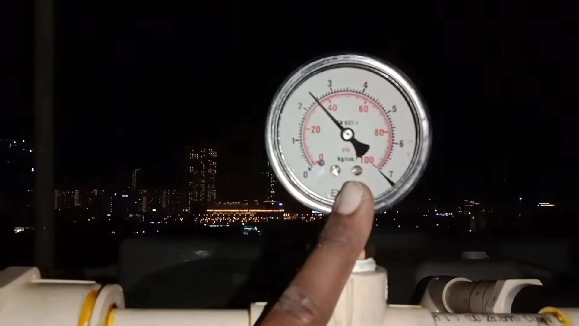 A finger pointing at a large round pressure gauge on a water line, with city lights blurred behind