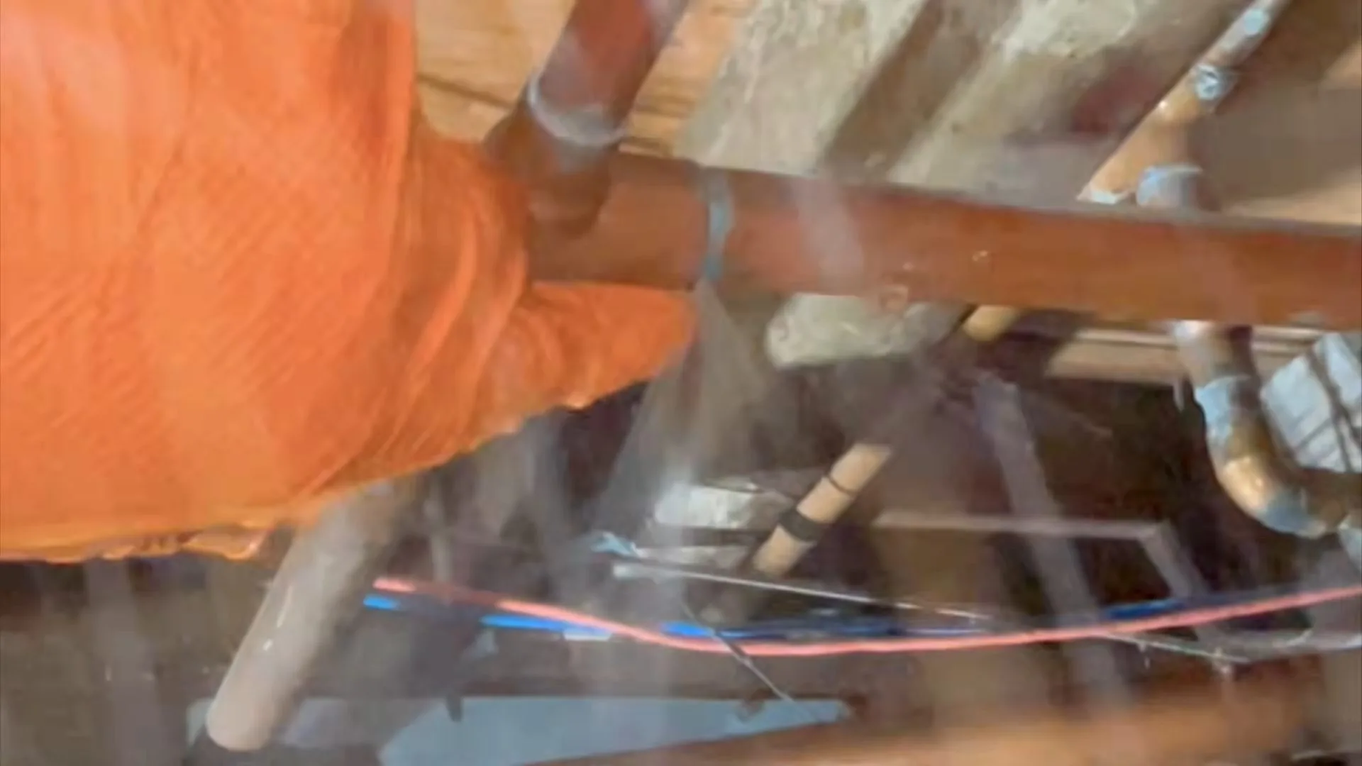 A gloved hand held against a copper pipe in a crawlspace while water sprays from it