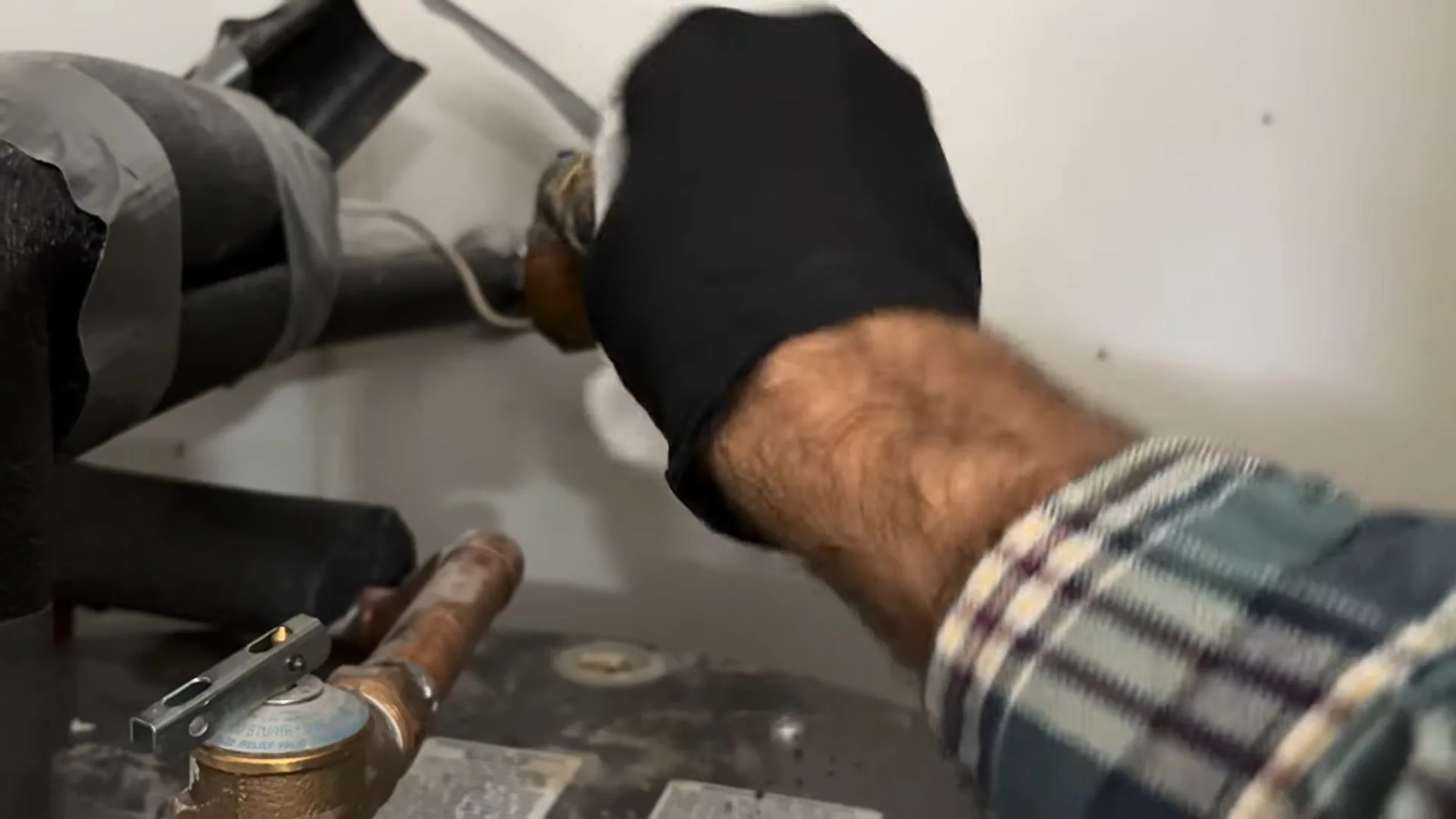 A gloved hand working on insulated pipes beside brass fittings above a water heater