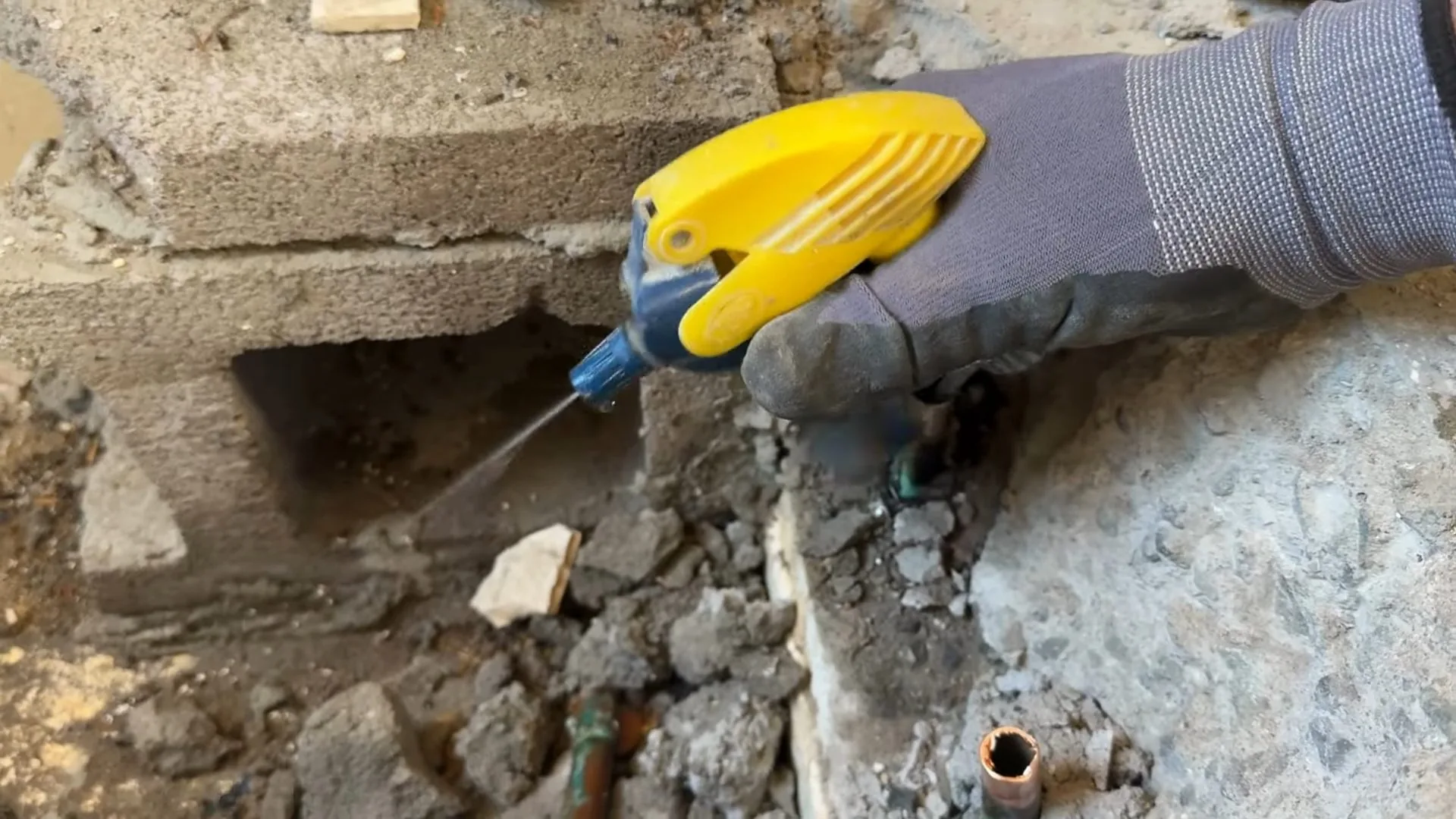 A gloved hand using a yellow handheld tool over broken concrete and block with a copper pipe end showing