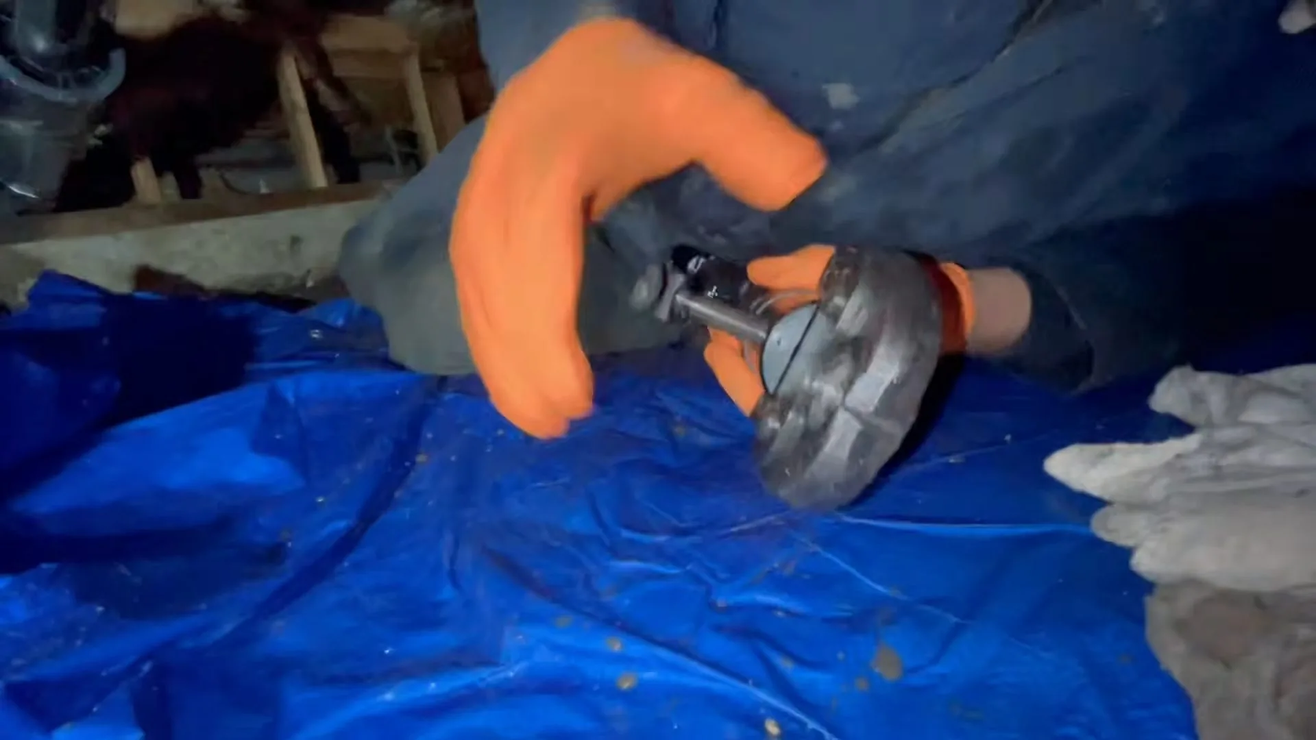 Hands in orange gloves holding a pipe fitting over a blue tarp in a crawlspace