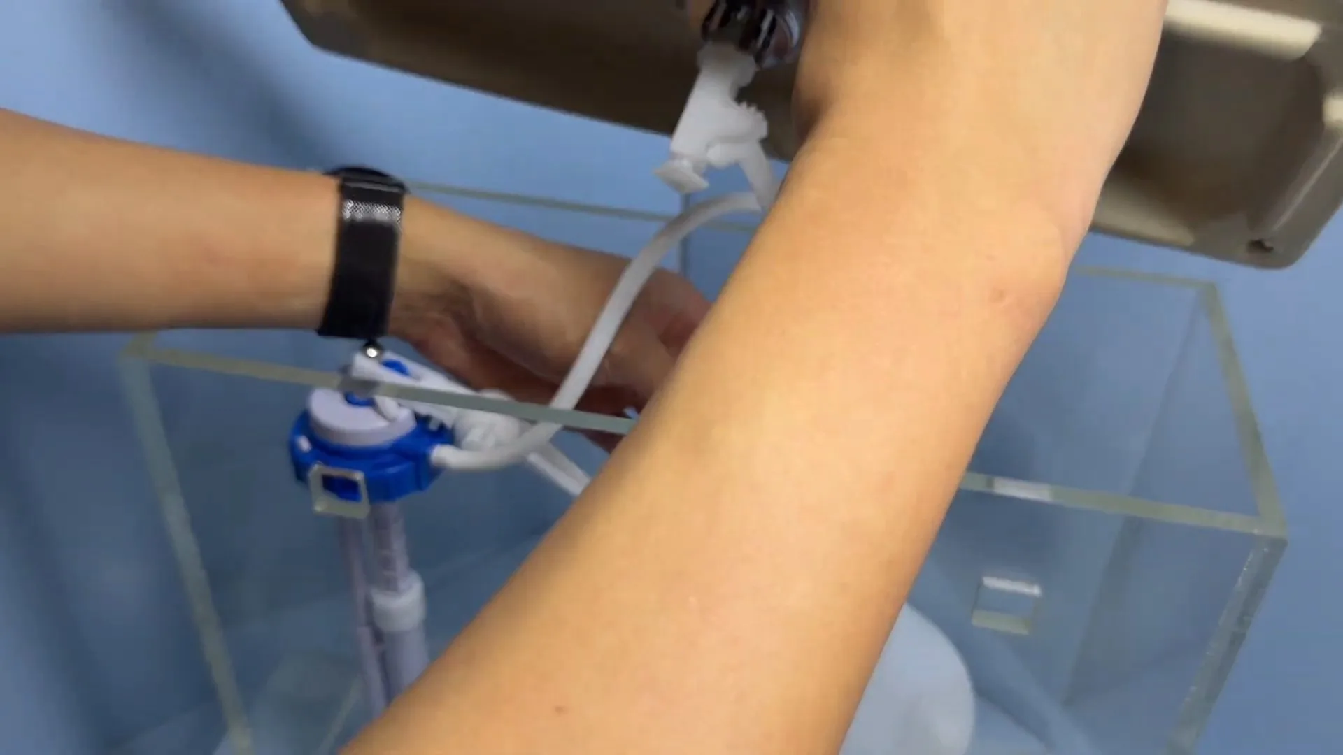 A hand adjusting a blue fill valve inside a clear acrylic toilet tank