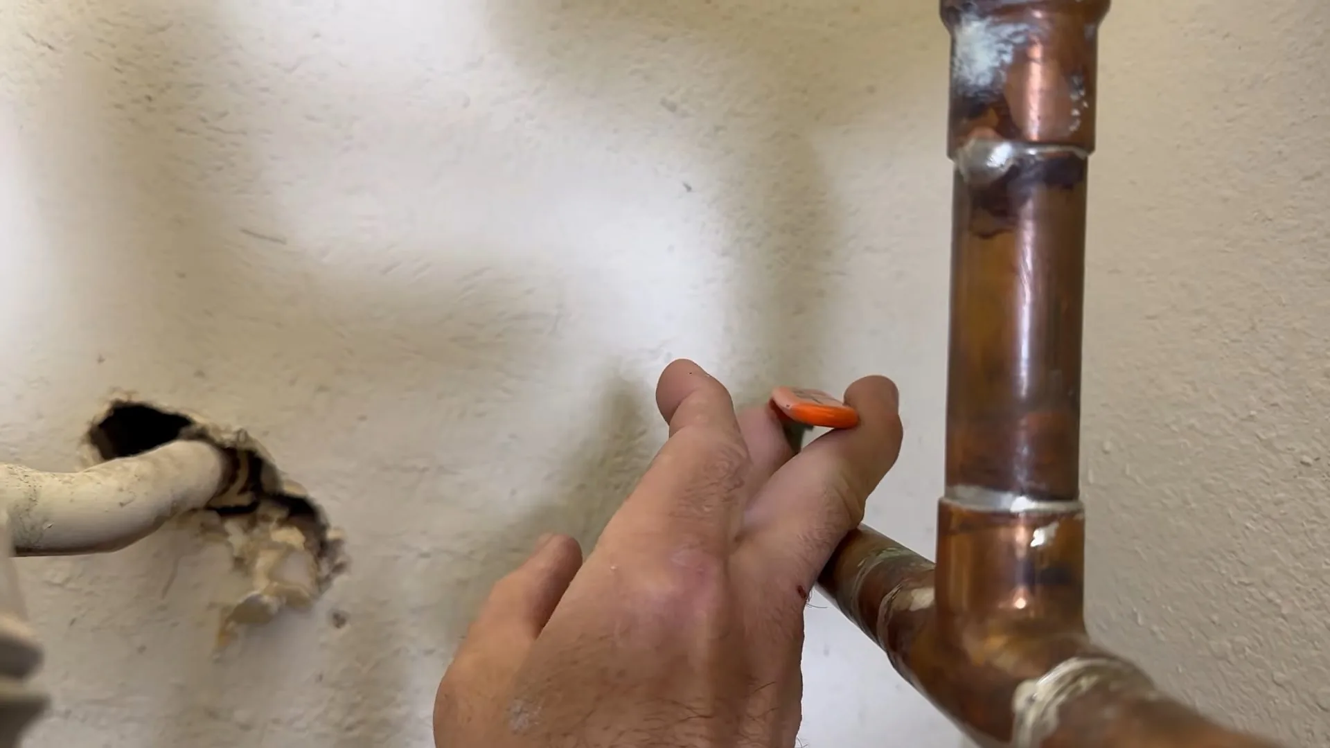A hand holding a small tool beside a copper pipe elbow with open holes in a white wall