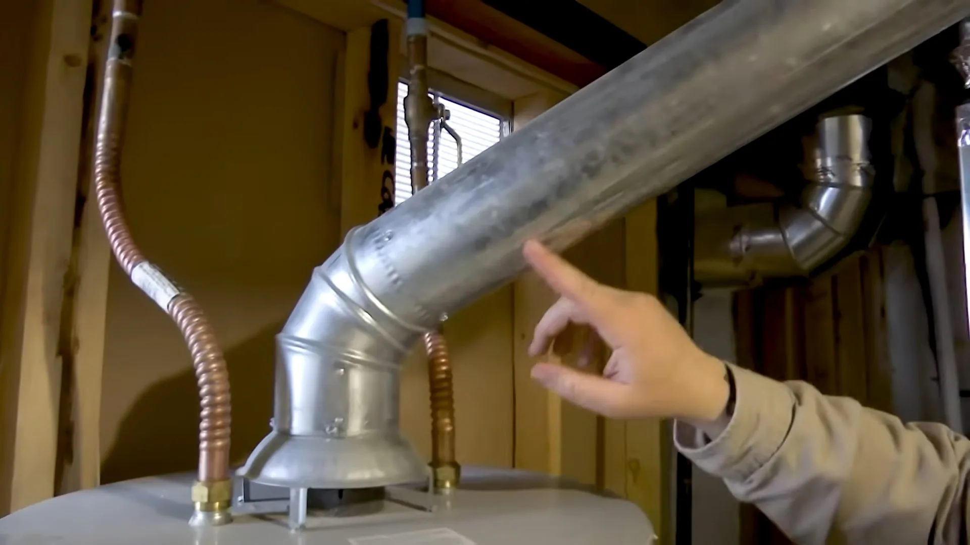 A hand pointing at a silver metal vent elbow on top of a water heater with flexible copper lines on either side