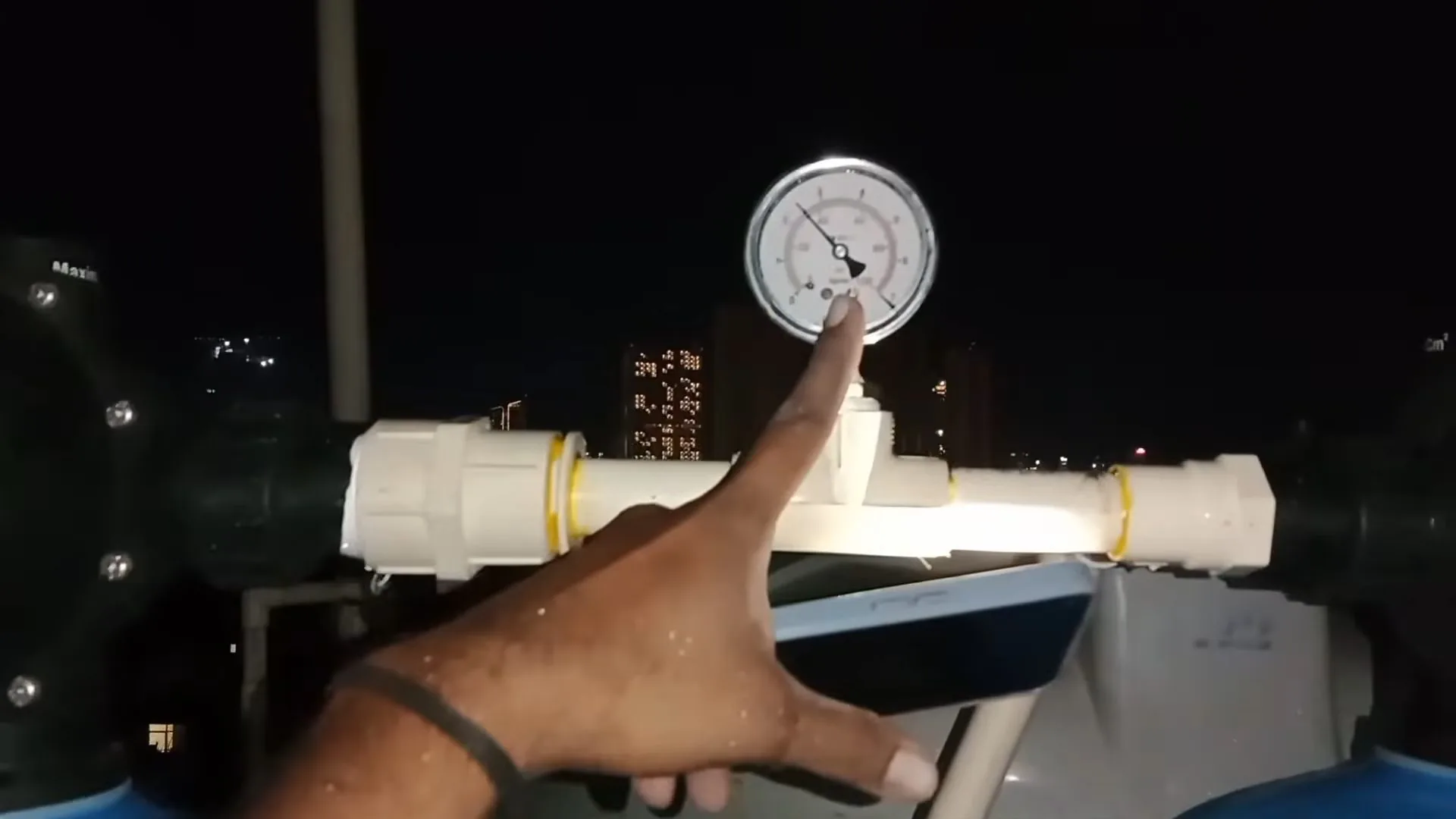 A hand pointing at a round pressure gauge mounted on a white water pipe at night