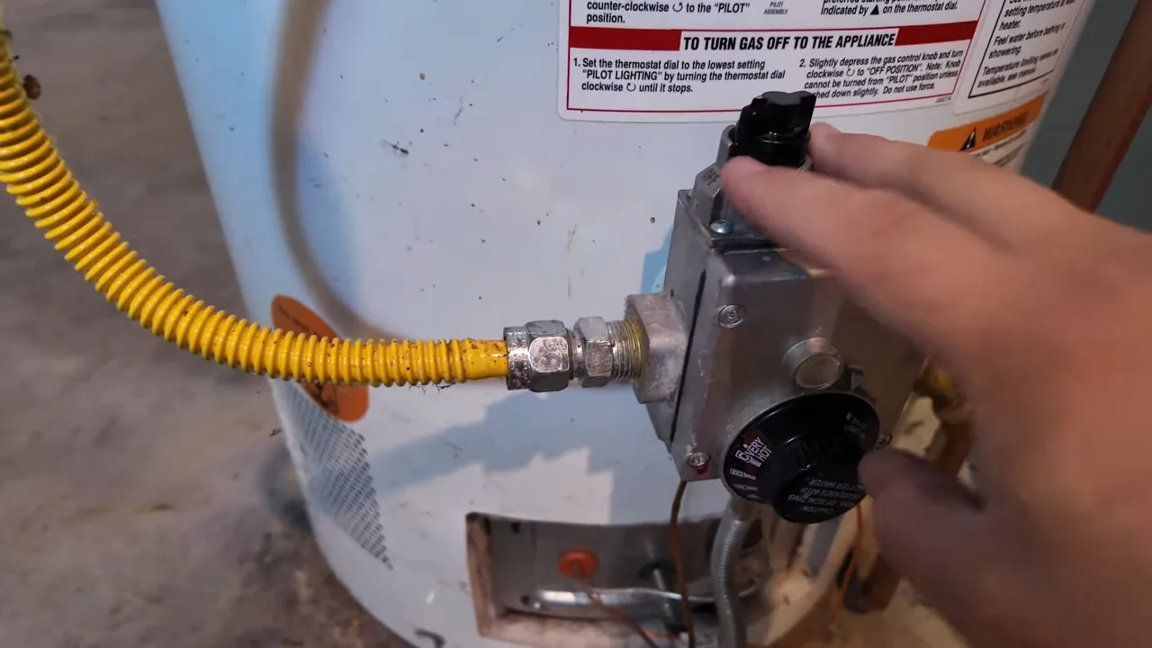 A hand turning the control knob on a water heater gas valve fed by a yellow flexible gas line