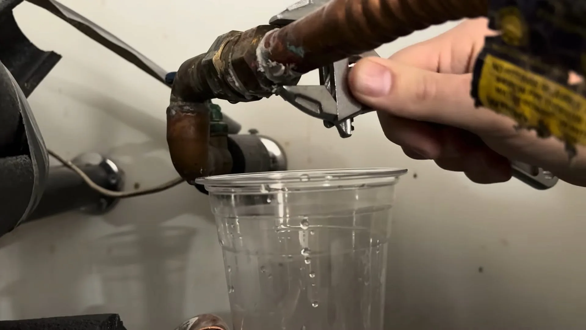 A hand holding a small tool at a copper pipe fitting above a clear plastic cup