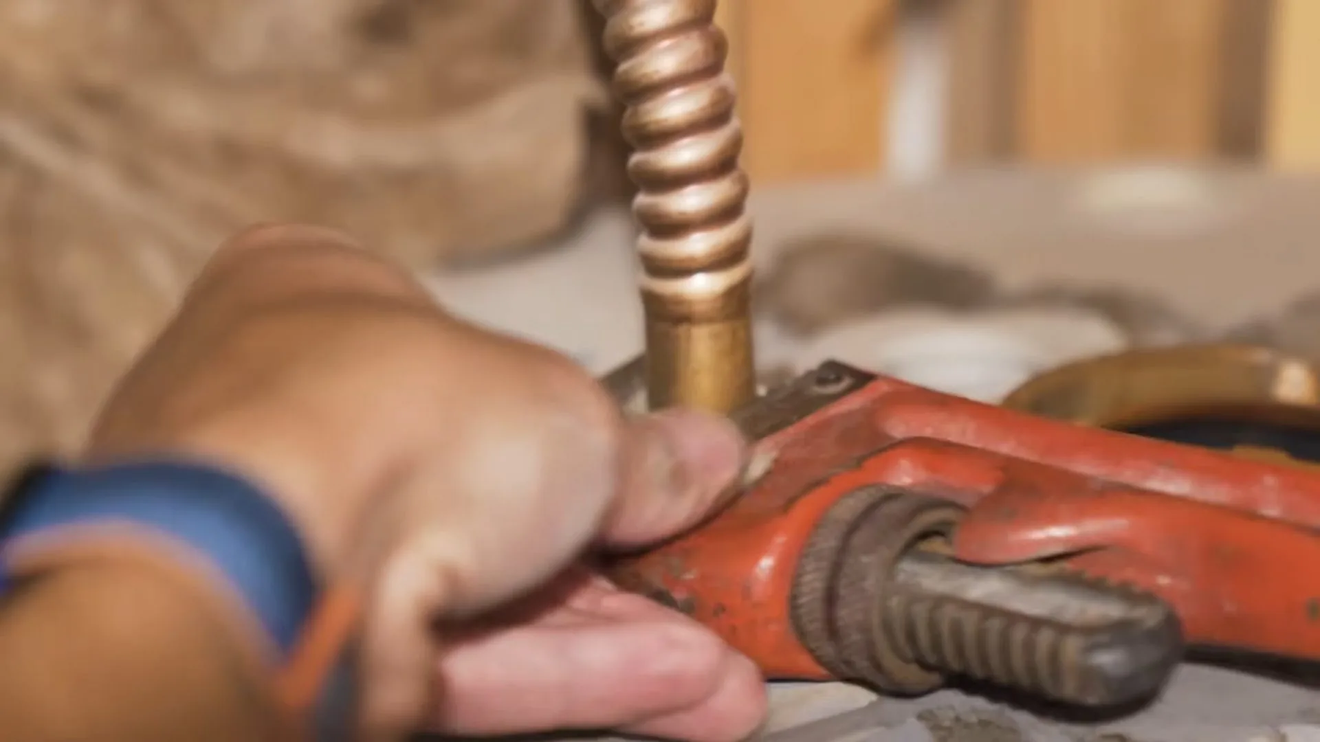 A hand gripping a red pipe wrench on a copper water line beside a flexible copper connector