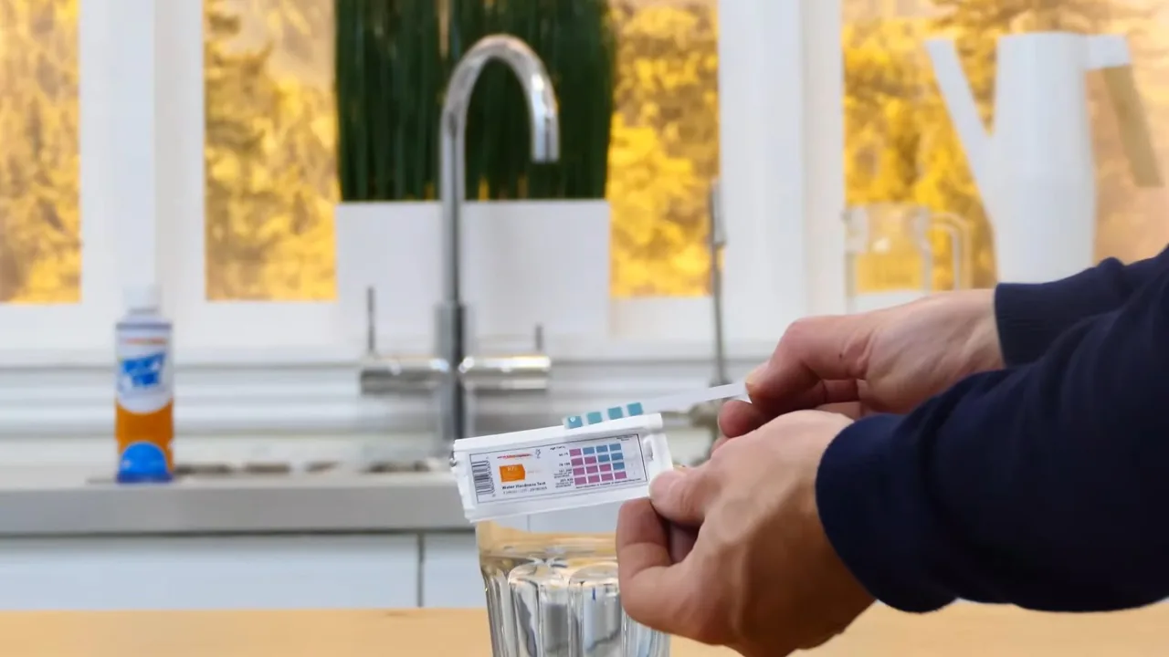 Hands holding a water test strip beside a kitchen tap and a drinking glass