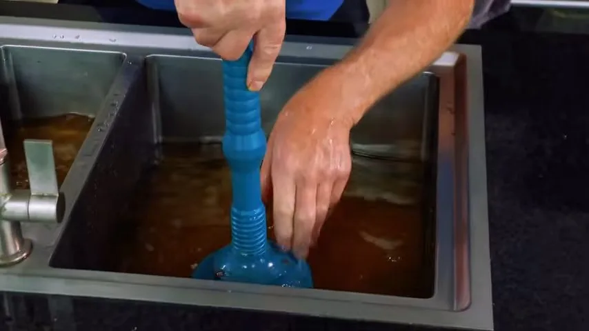 Two hands working a blue plunger in a double kitchen sink filled with brown water