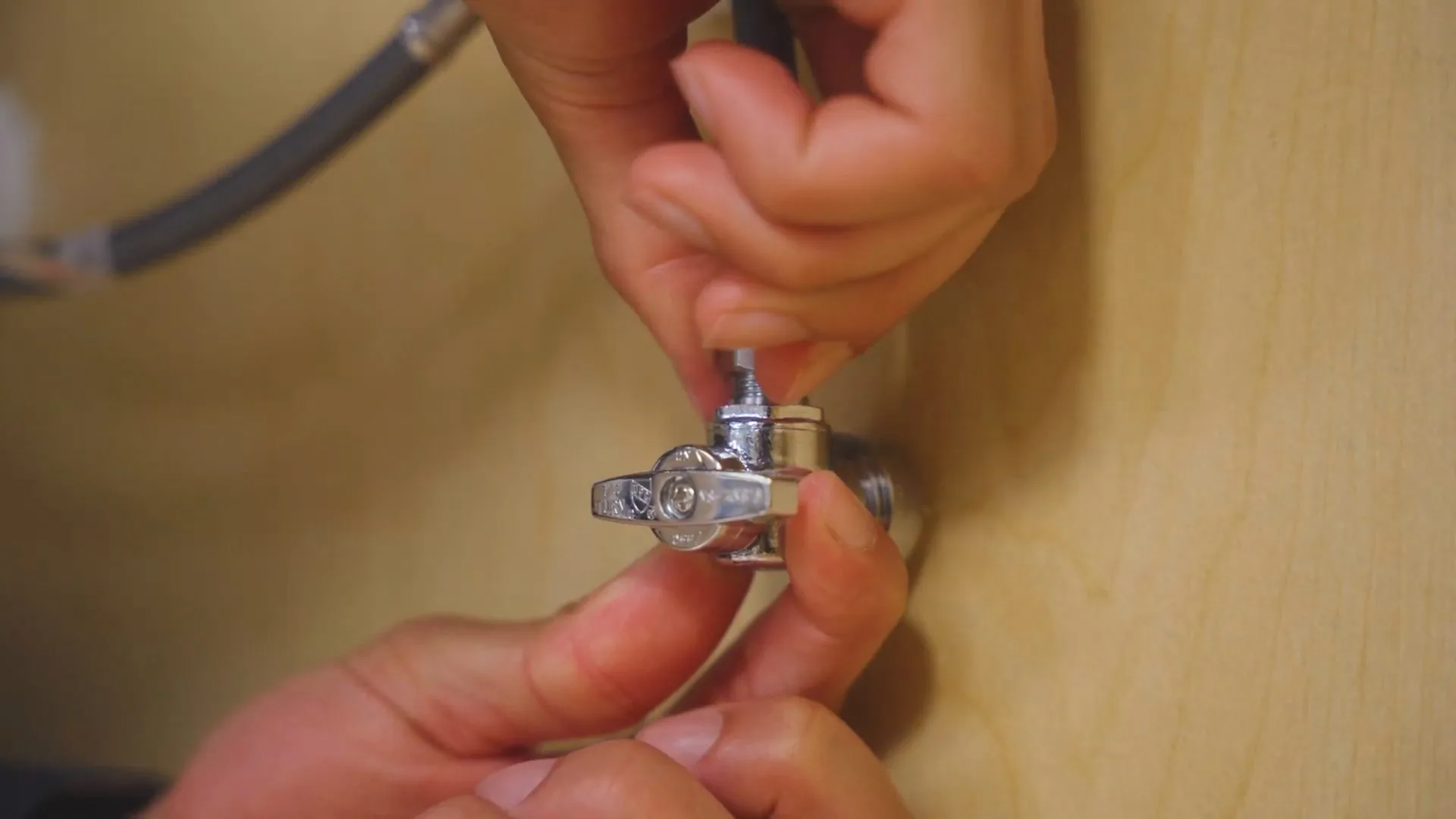 Hands turning the handle of a small chrome supply valve fitted to a flexible line against a wood wall