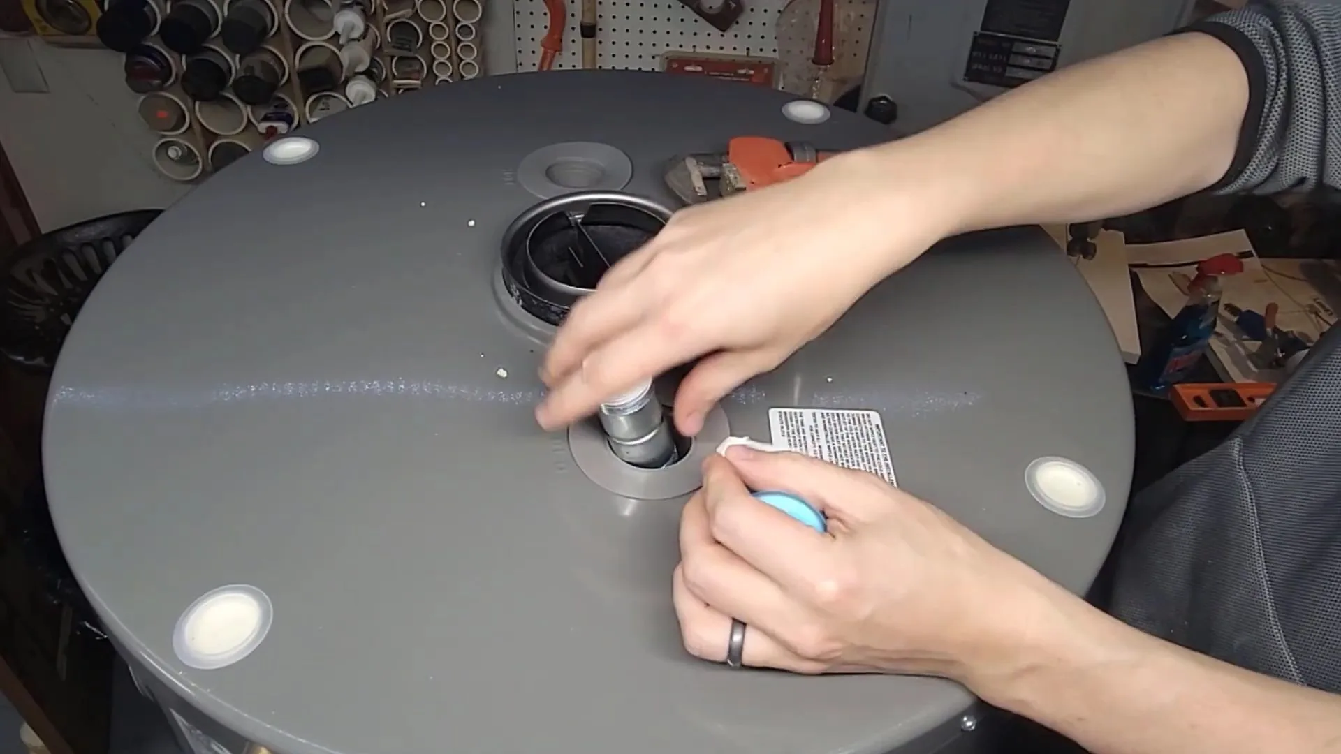Two hands working on a fitting at the top of a gray water heater tank in a workshop with pegboard behind