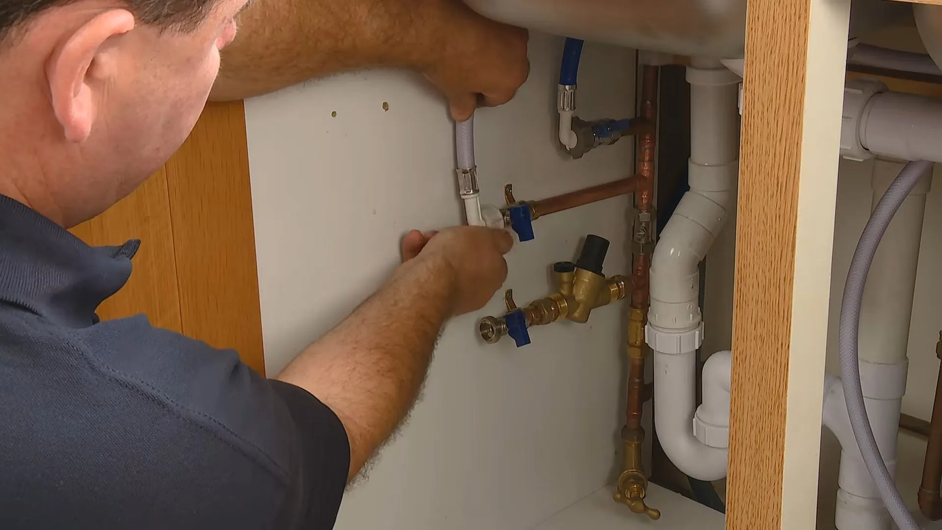 A man reaching into an open sink cabinet to work on blue-handled shutoff valves and white drain pipes