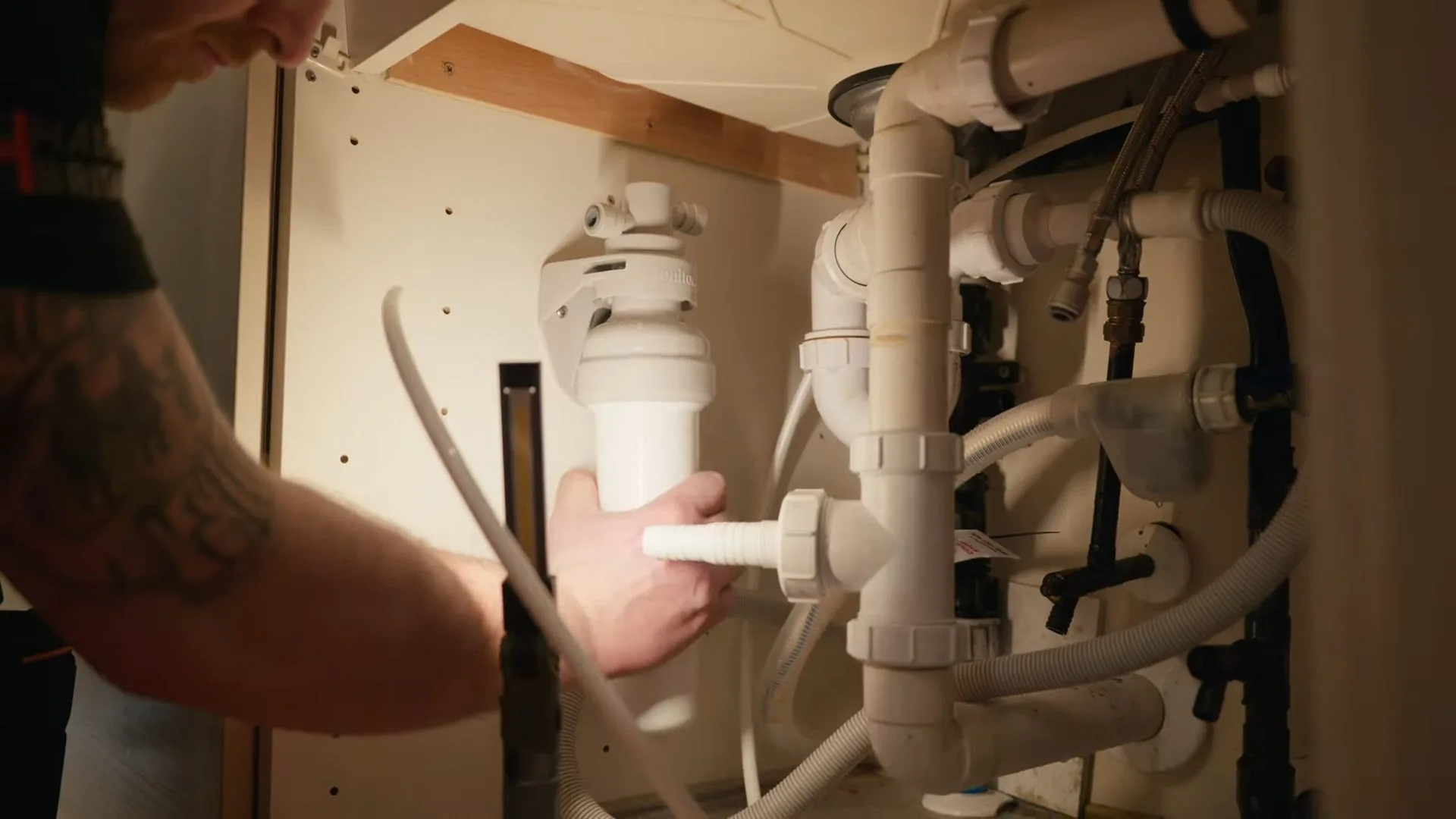 A man with a tattooed arm working on white pipes and tubing inside an under-sink cabinet