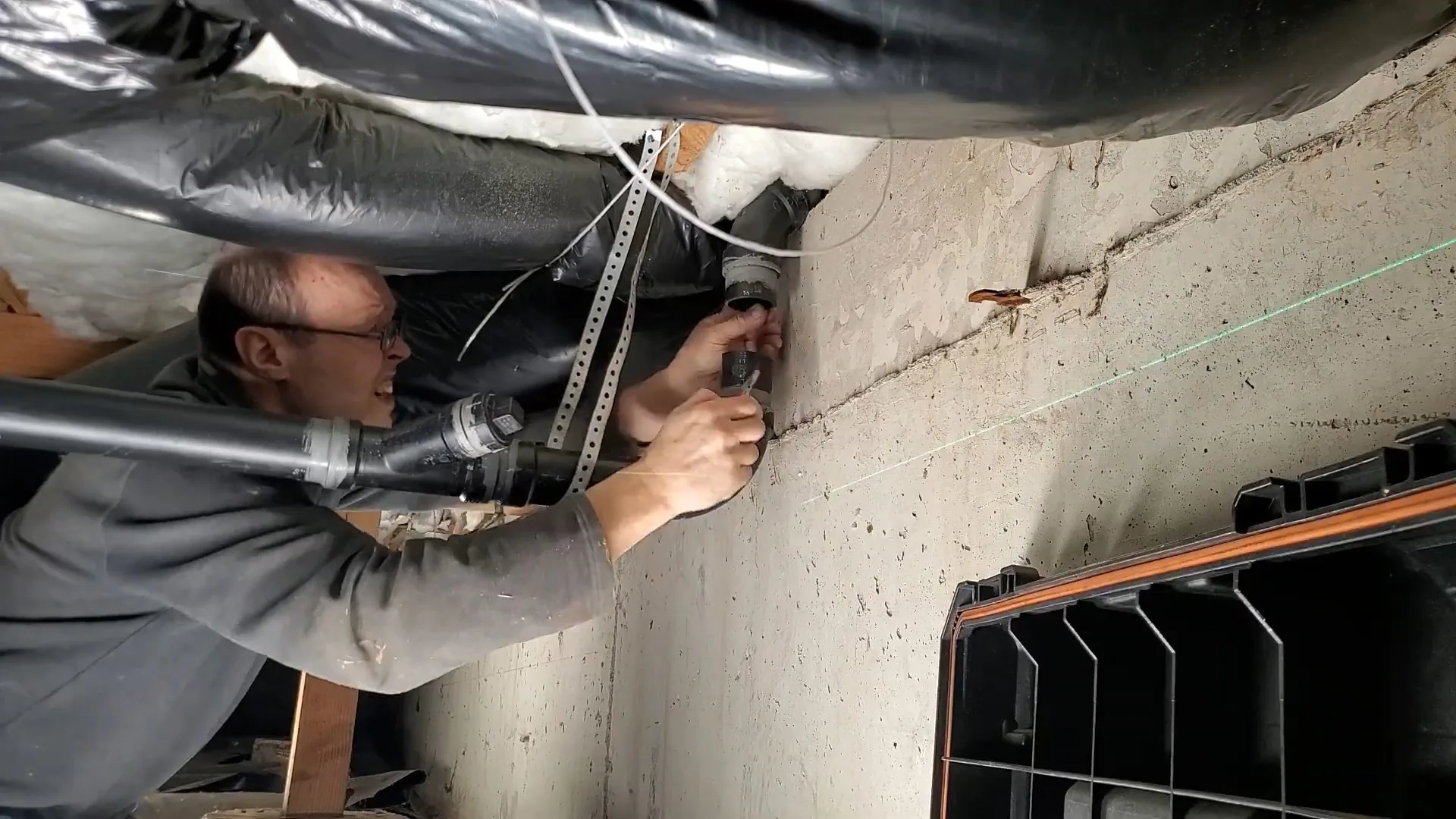 A man kneeling in a low crawlspace under ductwork, working on a black drain pipe beside a concrete wall with a green laser line
