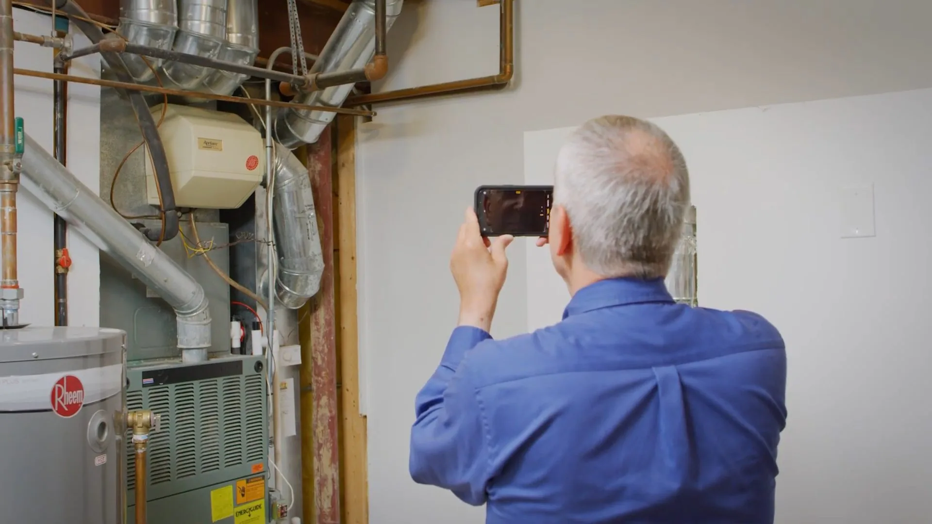 A man in a blue shirt photographing a utility room wall with a phone beside a water heater and furnace