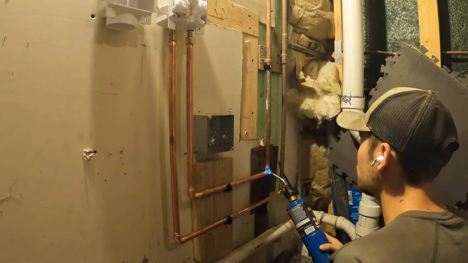 A man in a cap soldering copper pipes against an open wall in a basement with white plastic drain lines nearby