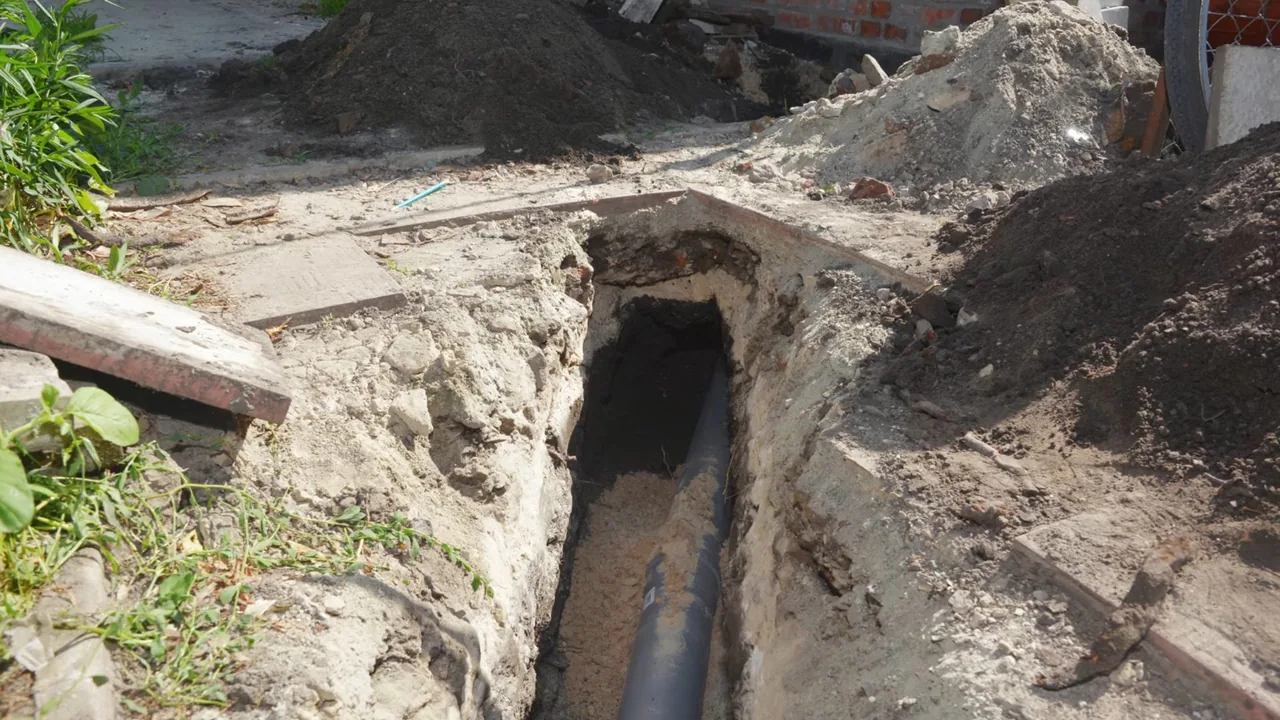 A narrow open trench beside a house with a black pipe laid in gravel at the bottom