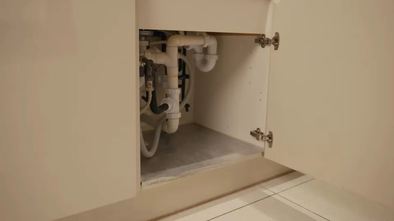 An open under-sink cabinet showing white drain traps and supply lines against the back wall