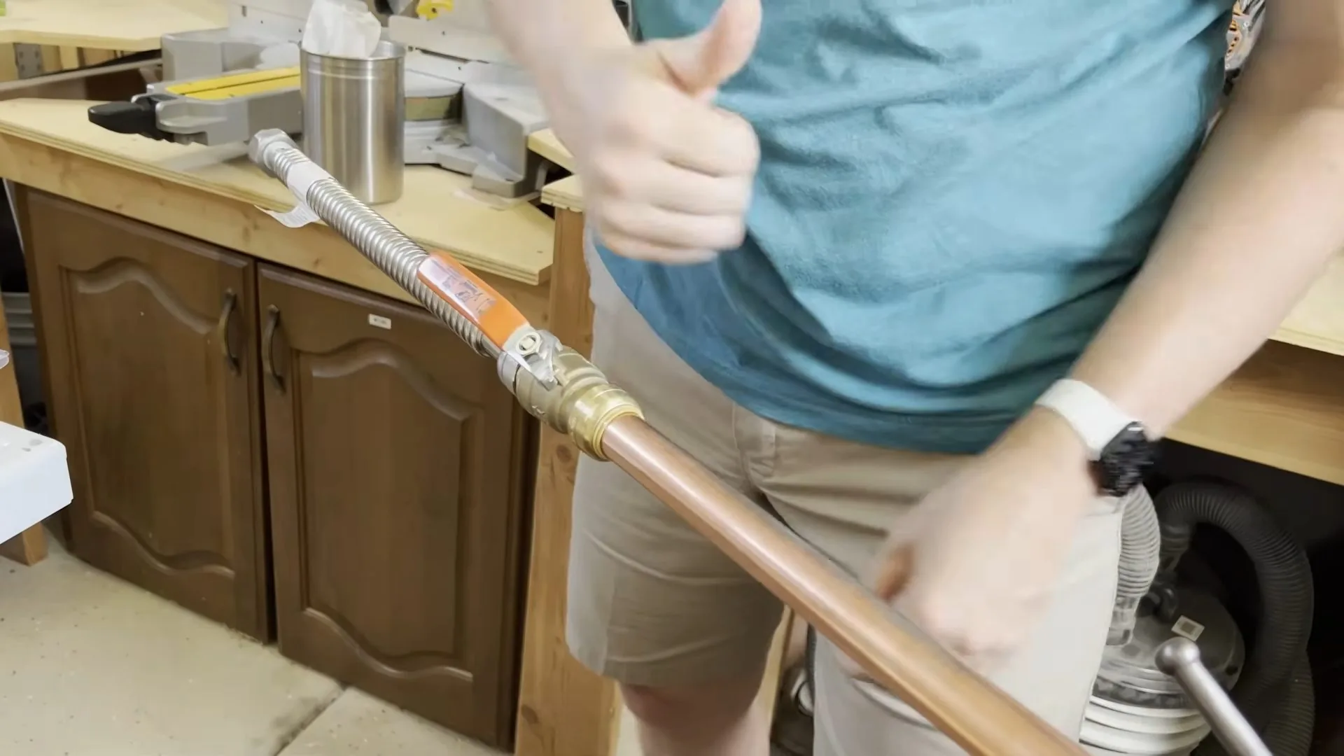 A person in a teal shirt giving a thumbs up while holding a copper pipe fitted with a valve beside wooden cabinets