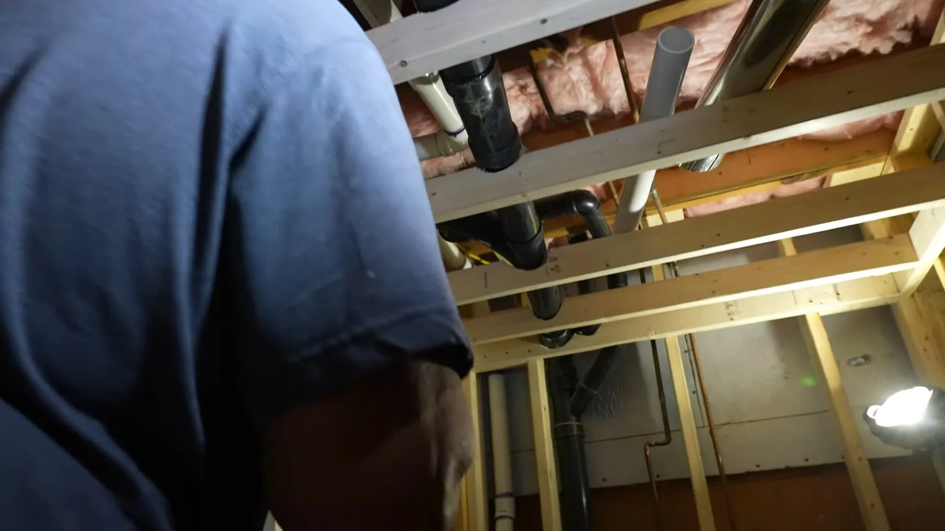 A plumber in a blue shirt working on black drain and vent piping between open basement joists