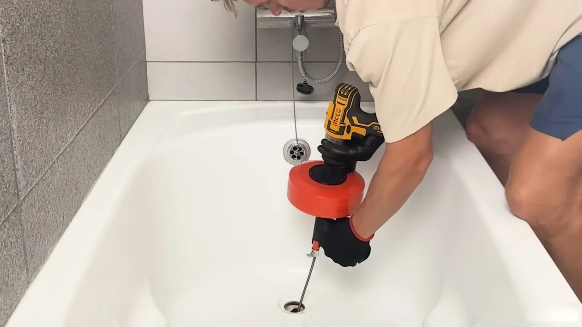 A person in a beige shirt using a cordless drill with a red drain auger in a white bathtub drain