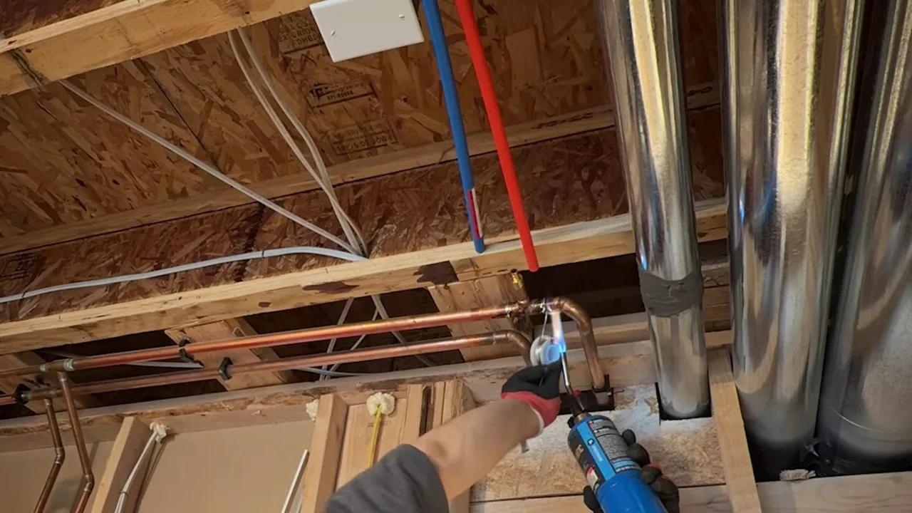 Red and blue plastic supply lines running along basement joists beside metal ductwork while a gloved hand holds a torch
