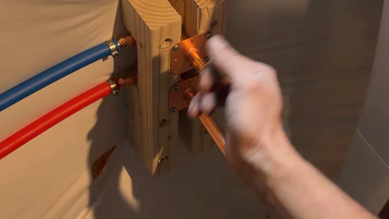 A hand holding a copper fitting beside red and blue flexible tubing clamped to copper stubs on a wooden frame