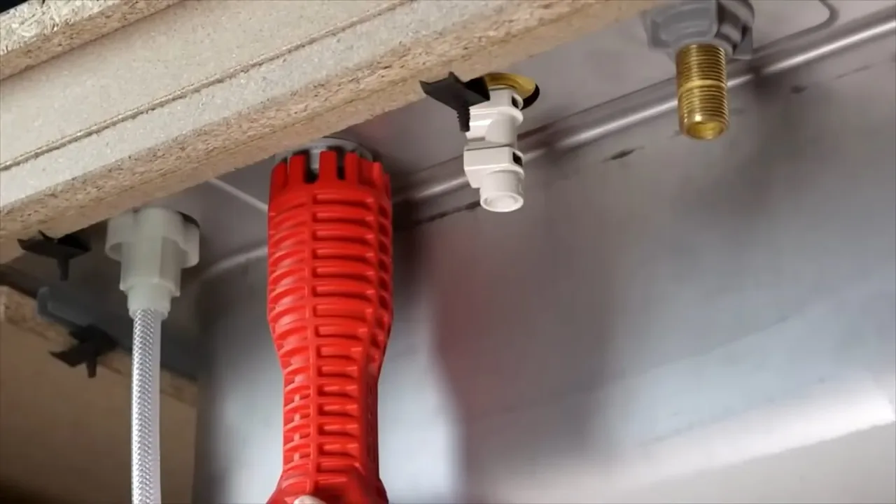 A red basin wrench resting under a sink beside a white drain connector and a braided supply line