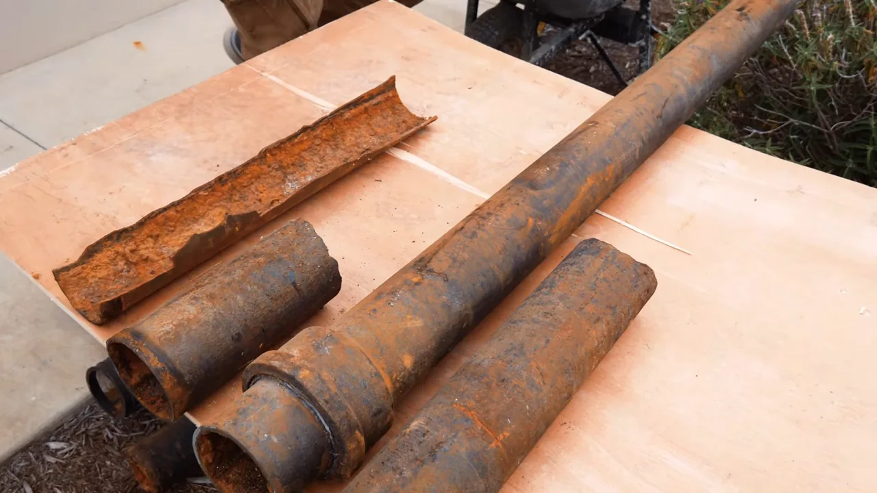 Several lengths of rusted cast iron pipe, one split open along its length, laid on a sheet of plywood outdoors