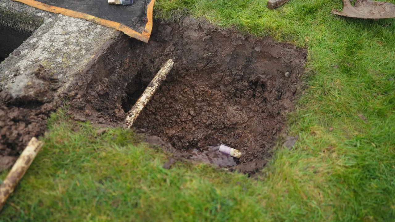 A small excavated hole in a lawn beside a concrete edge with a pipe end visible in the soil