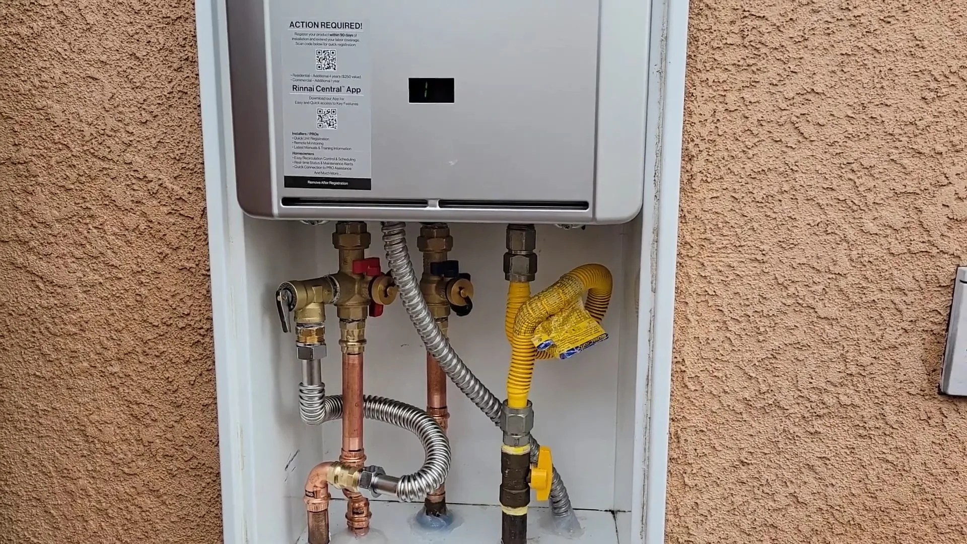 A white tankless water heater mounted in a recess of a stucco wall with gas and copper water lines below it