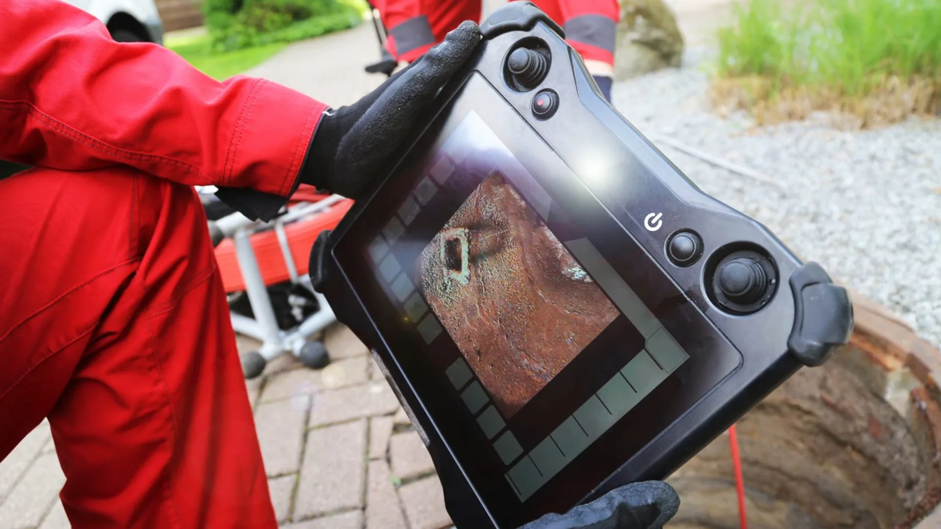 A technician in red coveralls holding a handheld camera monitor beside a round access opening on a paved path