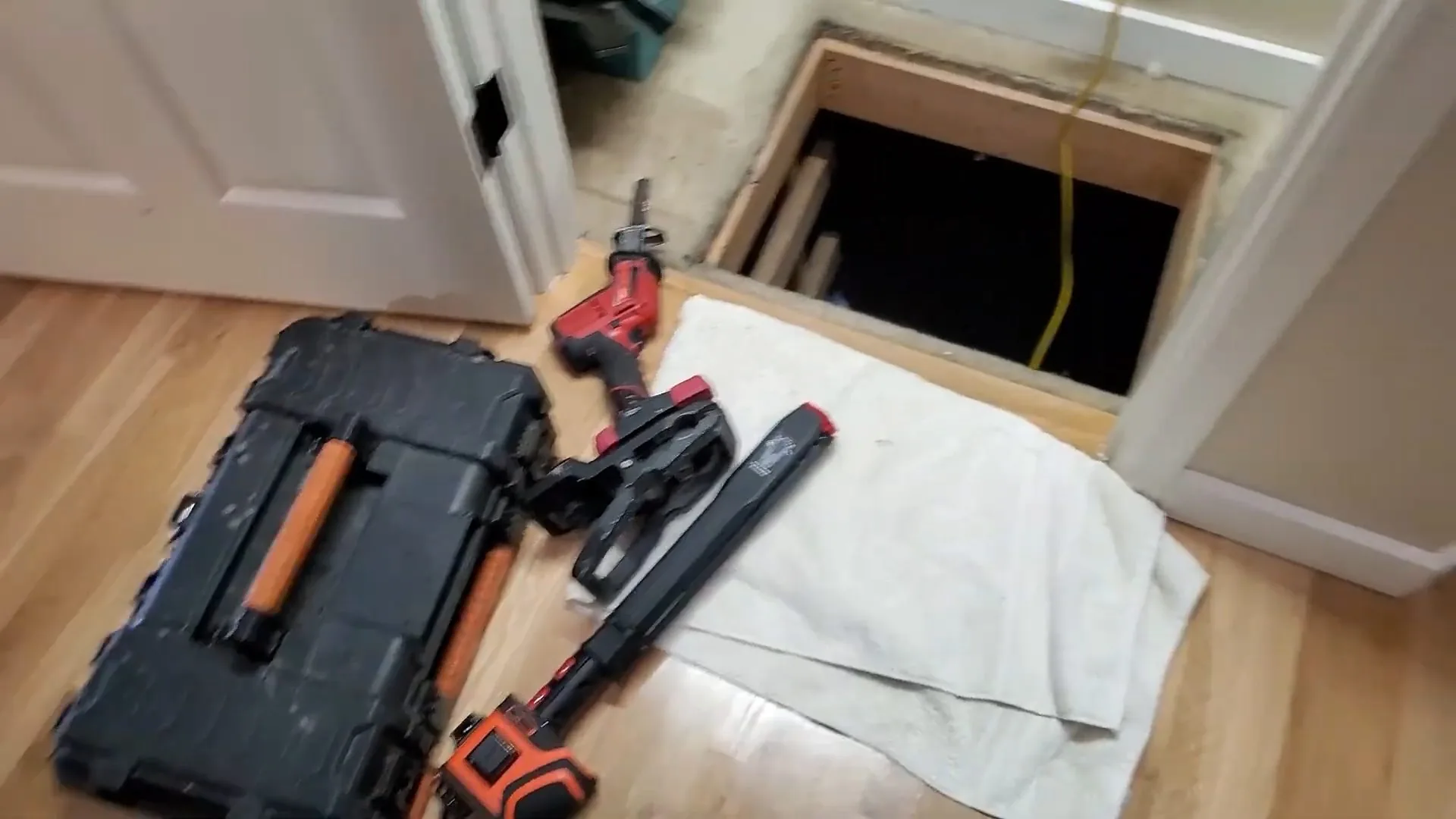 An open black tool case and two power tools on a towel beside an open floor access hatch