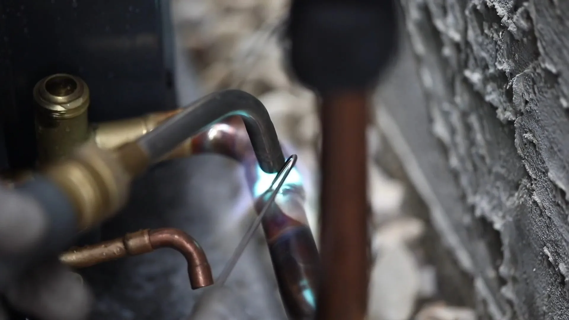 A torch flame heating a copper elbow joint beside a brass valve on a stucco wall
