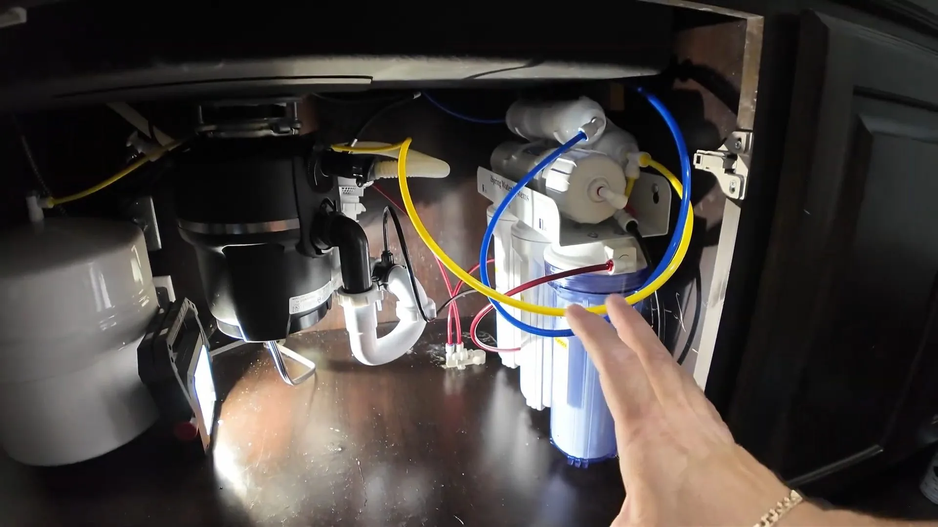 The inside of an under-sink cabinet showing a garbage disposal, white drain trap and blue and yellow filter tubing