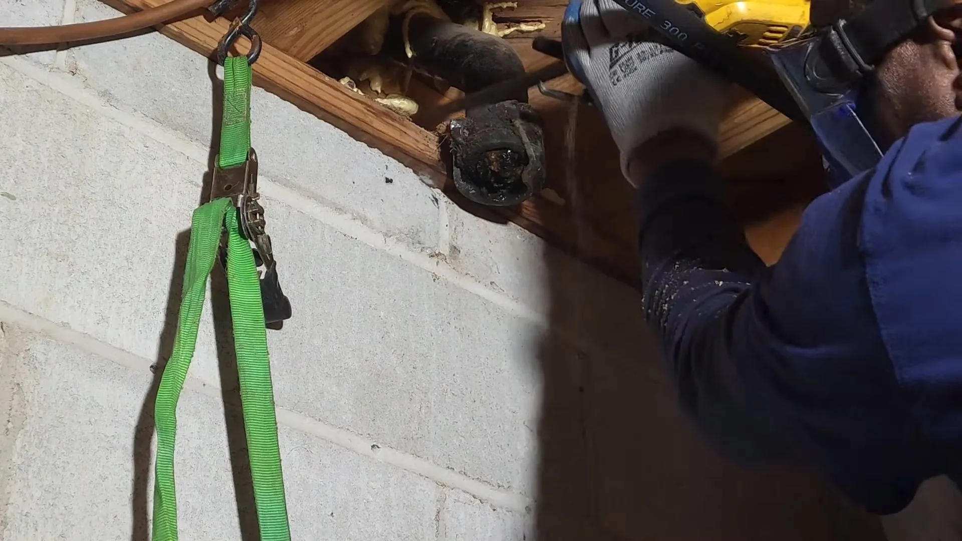 A plumber in a blue shirt using a yellow power tool to cut a cast iron pipe beneath wood joists, with a green strap hanging beside it