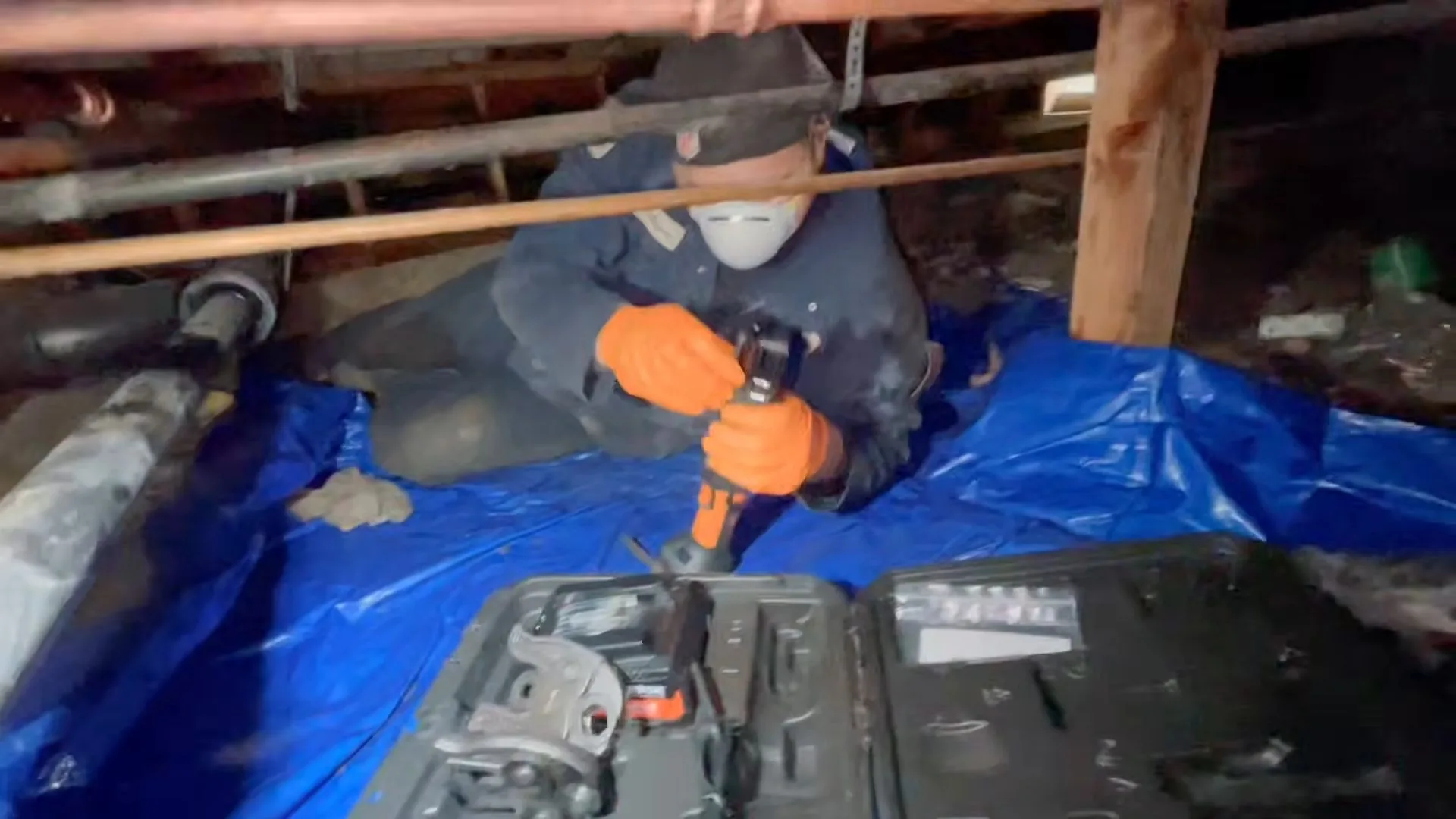A worker in a dust mask and orange gloves using a drill in a crawlspace on a blue tarp with an open tool case in front
