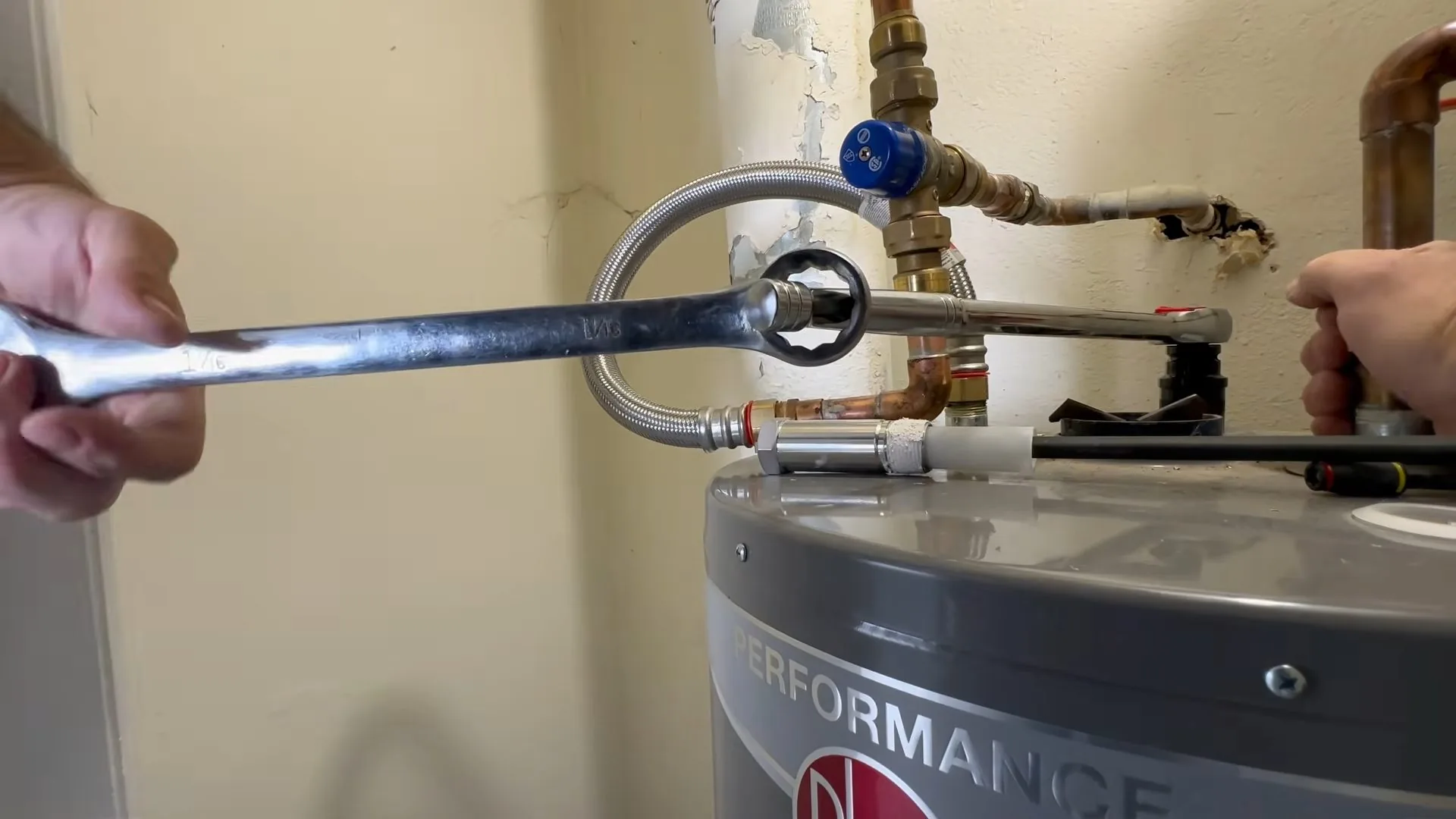 A hand holding a wrench on a flexible supply line above a gray water heater tank
